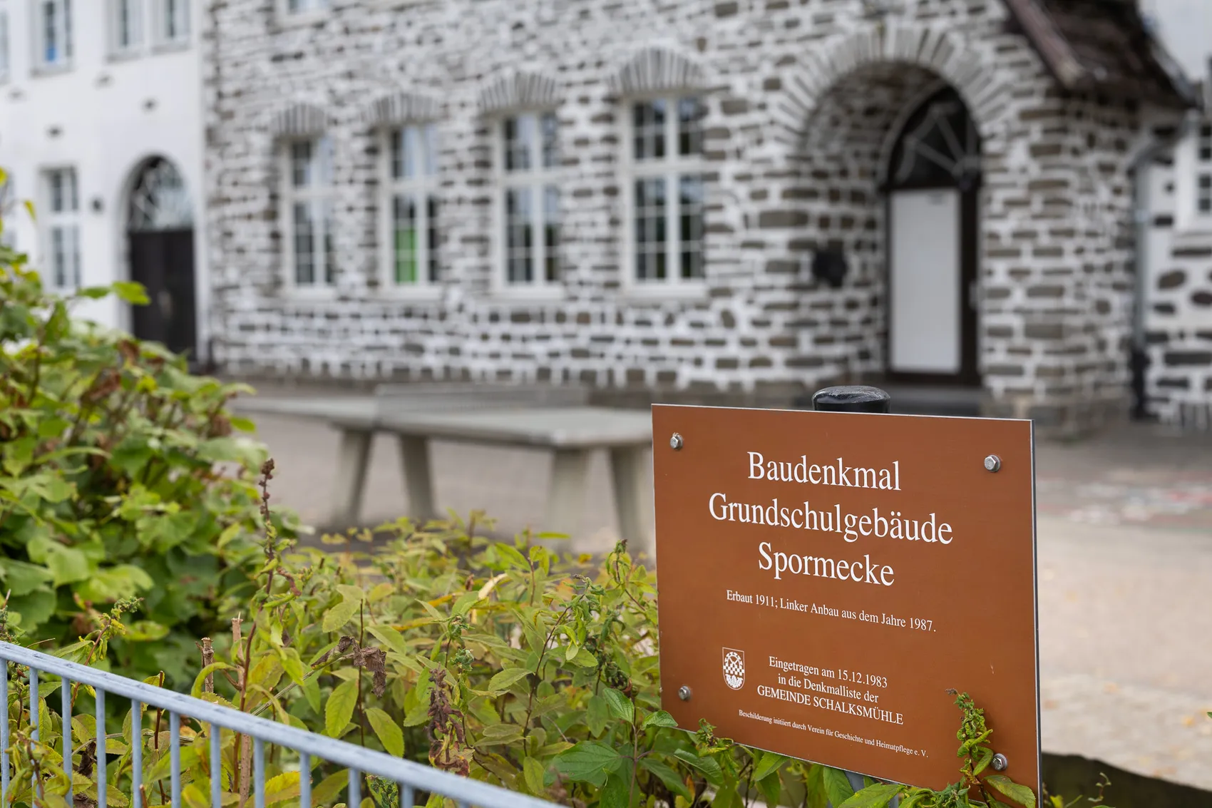 Braunes Hinweisschild zum Baudenkmal Grundschulgebäude Spormecke vor dem steinernen Schulgebäude.