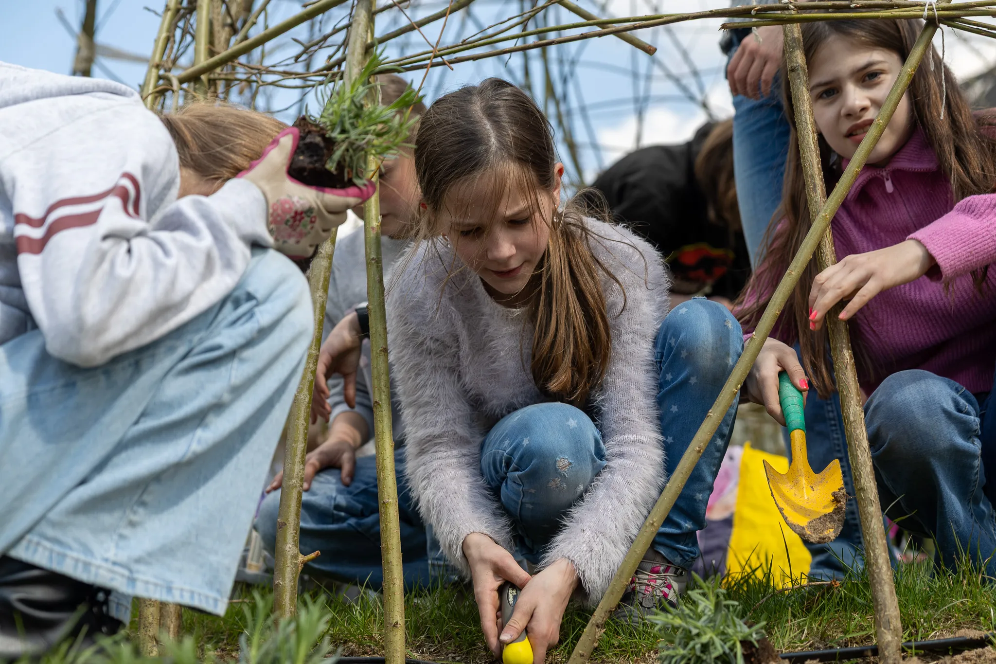 Schülerinnen knien im Garten und pflanzen Setzlinge. Eine von ihnen gießt die Pflanzen mit einer gelben Gießkanne.