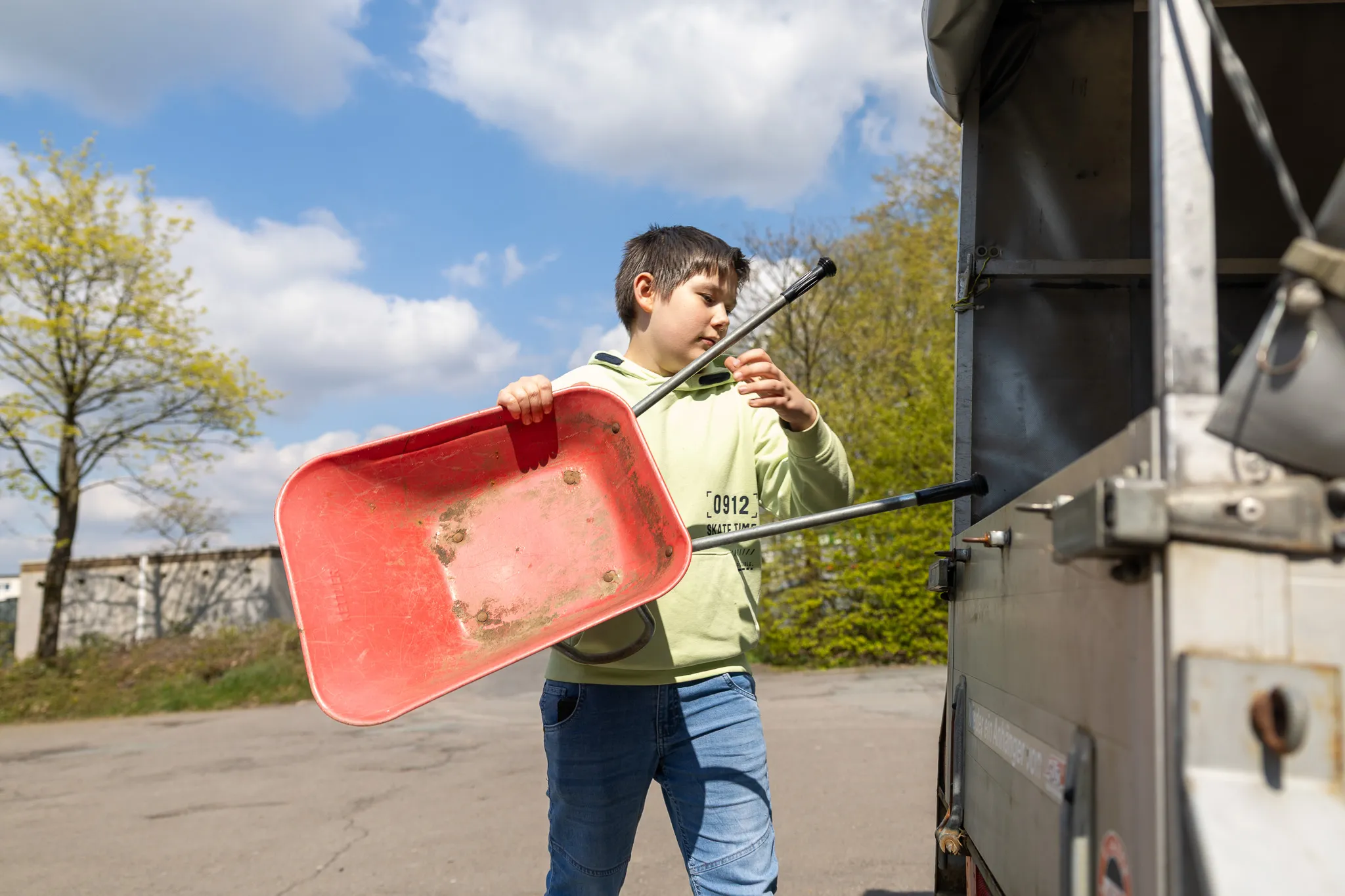 Ein Junge steht neben einem Transporter und hält eine rote Schaufel mit Stiel. Im Hintergrund sind Bäume und ein blauer Himmel zu sehen.