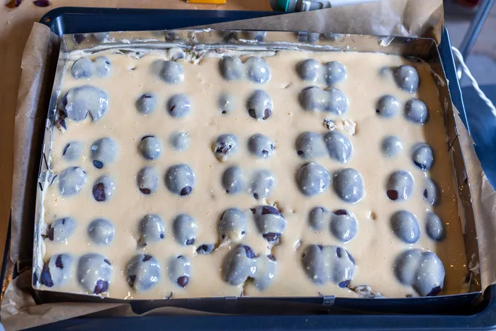 Heller Teig mit ganzen Pflaumen in einer mit Backpapier ausgelegten Backform.