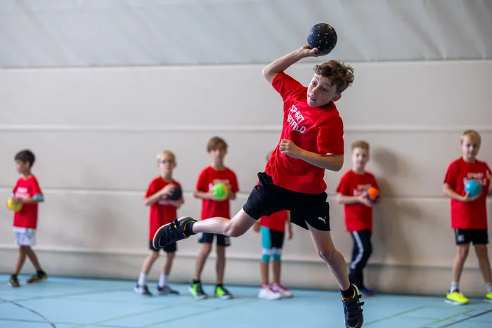 Ein Junge in einem roten T-Shirt springt in einer Sporthalle hoch und wirft einen dunklen Ball.