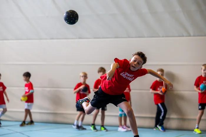 Ein Junge in einem roten T-Shirt springt in einer Sporthalle und wirft einen Ball, während andere Kinder im Hintergrund stehen.