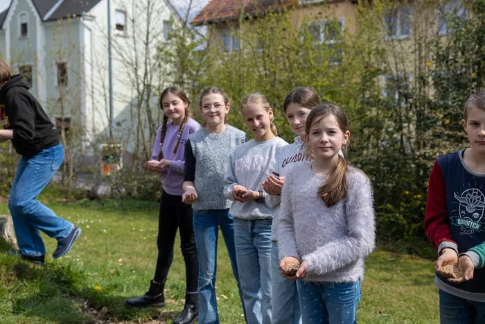 Eine Gruppe Jugendlicher steht auf einem Gehweg vor Häusern und hält kleine Säcke in den Händen, vermutlich für Samen oder Pflanzen.