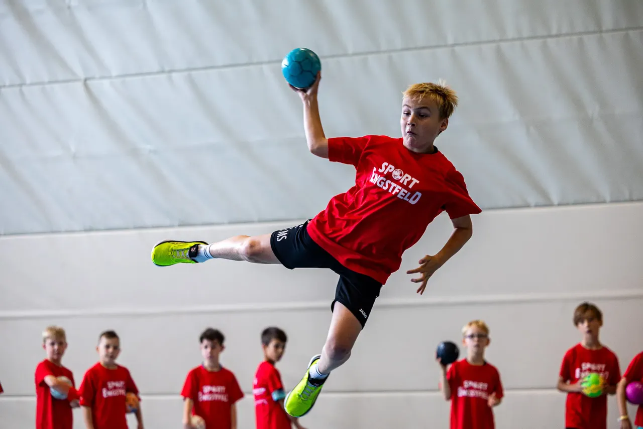 Ein Junge in einem roten T-Shirt springt mit einem blauen Ball in der Hand hoch, um ihn zu werfen.