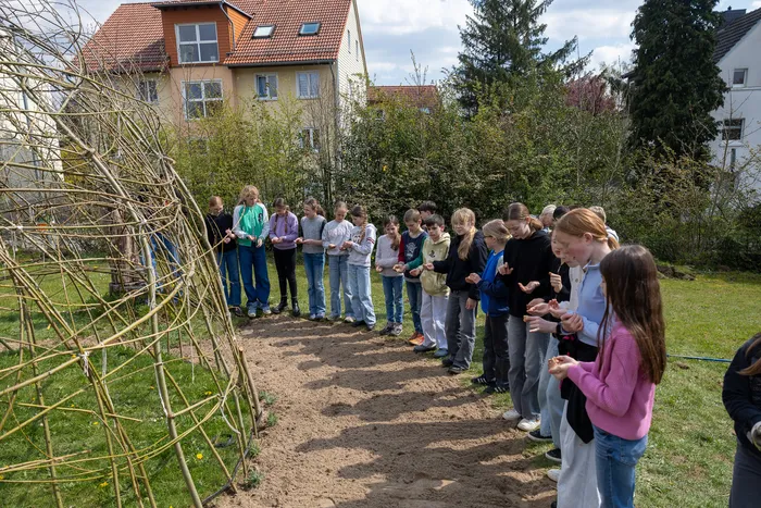 Eine Gruppe Jugendlicher steht im Garten und betrachtet einen aus Ästen geflochtenen Bogen. Im Hintergrund sind Häuser und Bäume zu sehen.