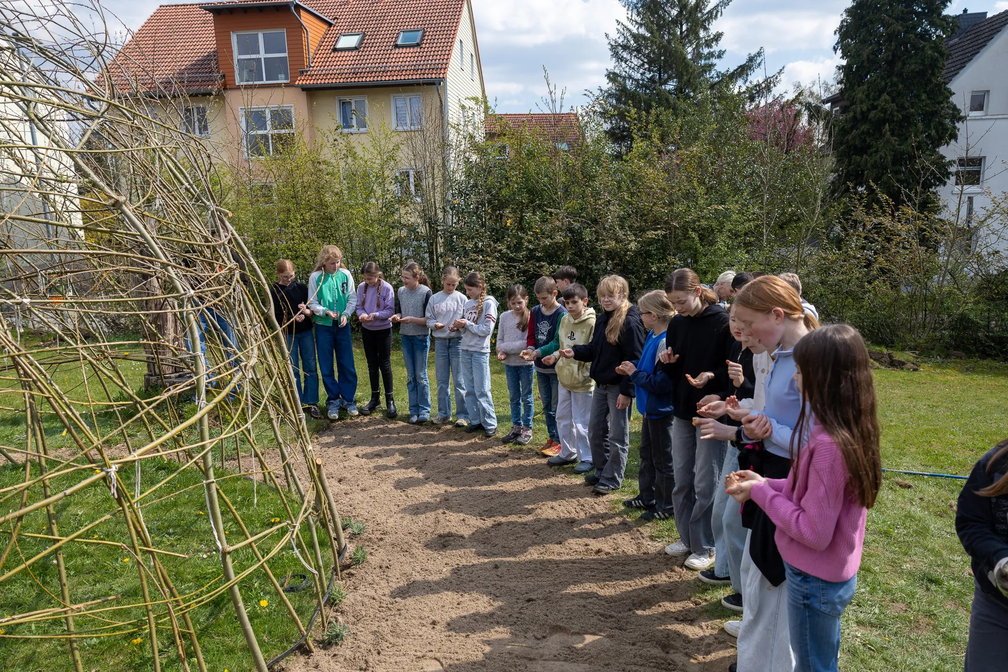 Eine Gruppe Jugendlicher steht im Garten und betrachtet einen aus Ästen geflochtenen Bogen. Im Hintergrund sind Häuser und Bäume zu sehen.