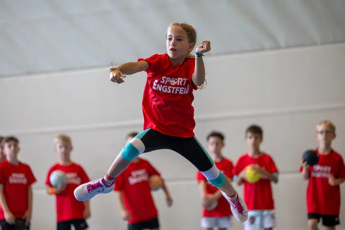 Ein Mädchen in einem roten T-Shirt mit der Aufschrift „SPORT ENGSTFELD“ springt in einer Sporthalle hoch in die Luft.