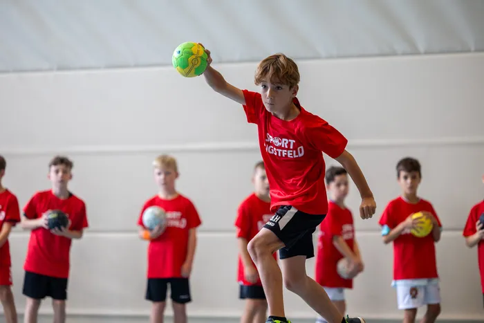 Ein Junge in einem roten T-Shirt wirft einen gelb-grünen Ball, während andere Kinder in roten T-Shirts im Hintergrund stehen.