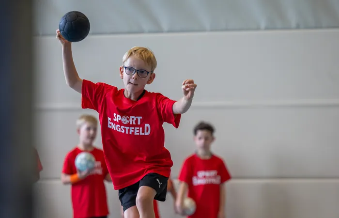 Ein Junge mit Brille und rotem T-Shirt mit der Aufschrift „SPORT ENGSTFELD“ hält einen schwarzen Ball zum Wurf bereit.