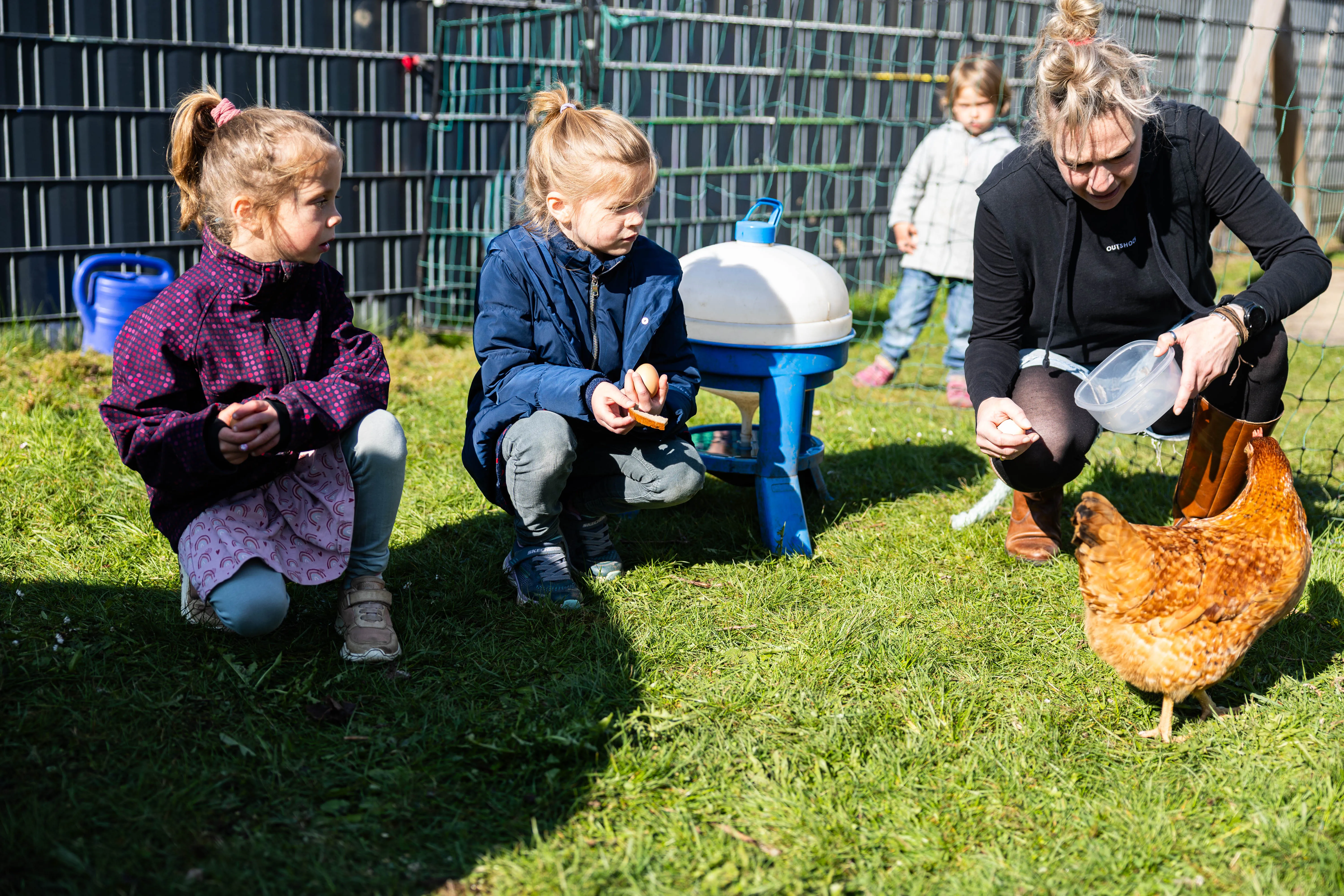 Drei Personen – zwei Kinder und eine Frau – hocken auf einer Wiese und füttern Hühner. Im Hintergrund sind weitere Kinder und ein Zaun zu sehen.