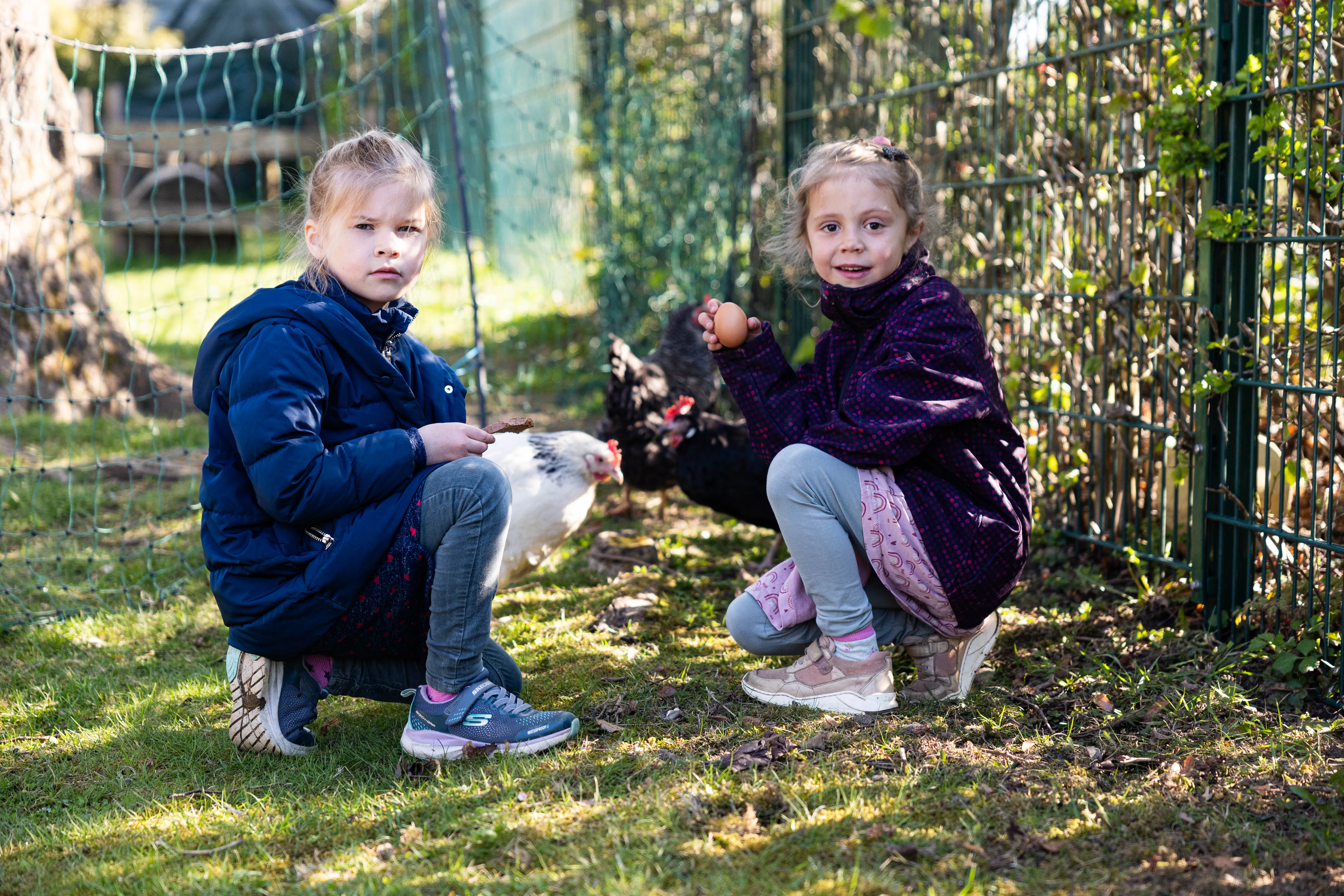 Zwei kleine Mädchen kauern im Gras vor einem Zaun und halten Hühnerfutter bzw. ein Ei. Im Hintergrund sind weitere Hühner und Büsche zu sehen.