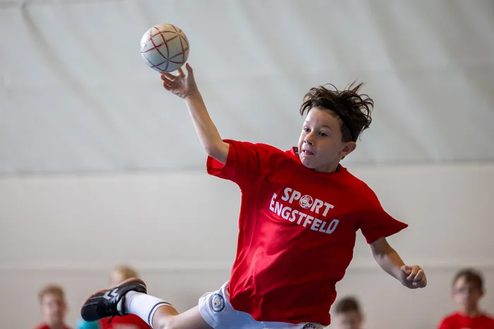 Ein Junge in einem roten T-Shirt mit der Aufschrift „SPORT ENGSTFELD“ springt und wirft einen weißen Ball mit roten Linien.