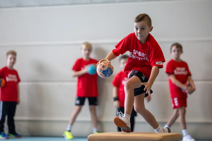Ein Junge in einem roten T-Shirt springt mit einem Handball auf eine Turnbank in einer Sporthalle.