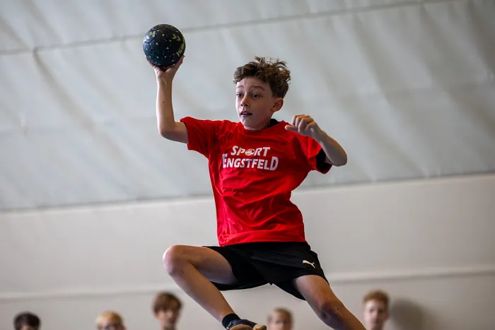 Ein Junge in einem roten T-Shirt mit der Aufschrift „SPORT ENGSTFELD“ springt mit einem Ball in der erhobenen Hand.