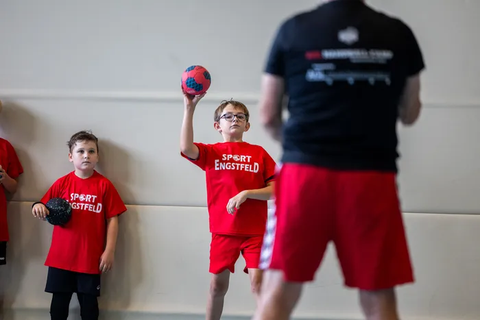 Ein Junge in einem roten „Sport Engstfeld“-T-Shirt hält einen rot-blauen Ball hoch, während ein weiterer Junge daneben einen schwarzen Ball hält.