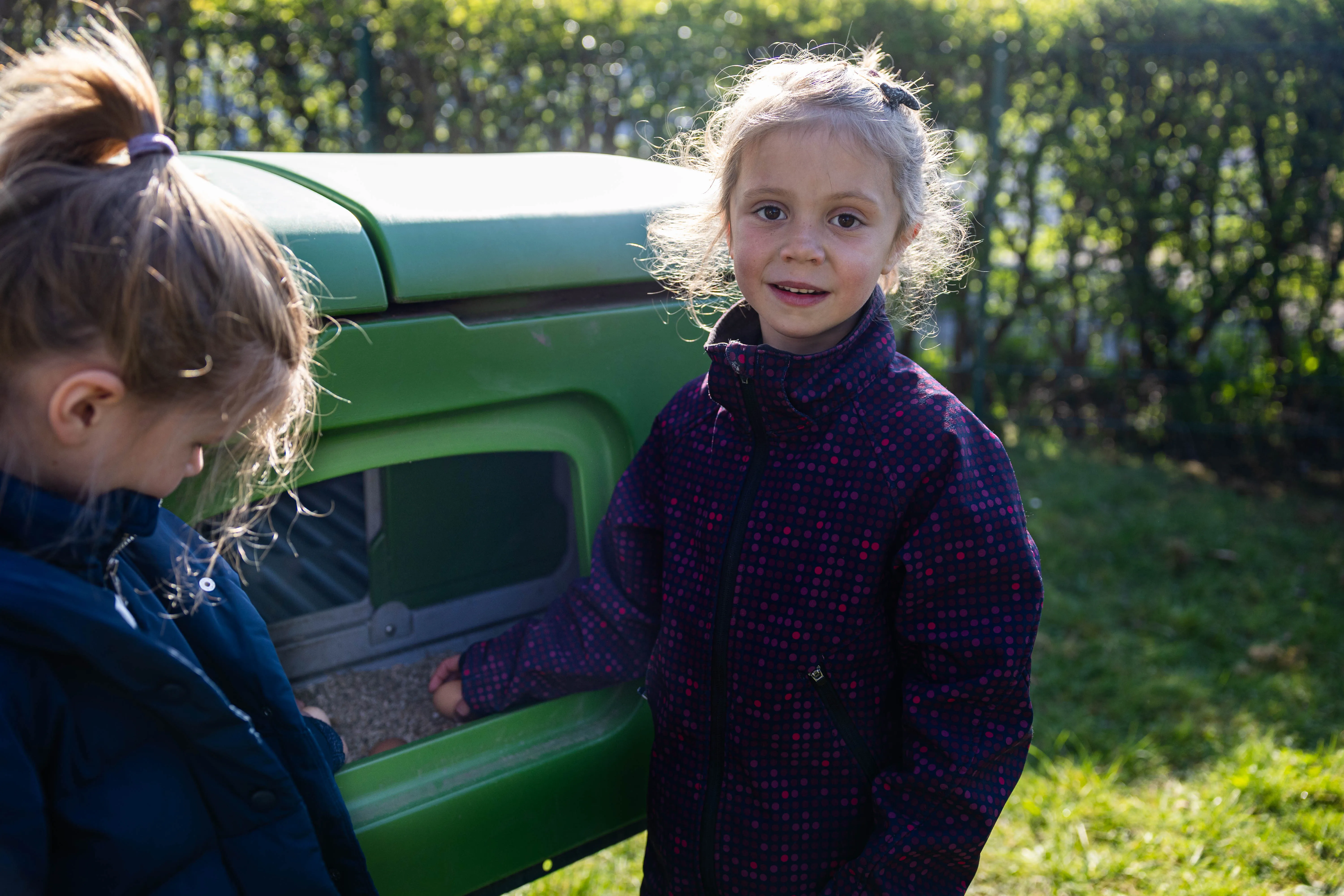 Zwei Mädchen betrachten Eier in einem grünen Hühnerstall. Charlotte blickt in die Kamera, während Sophie sich dem Stall zuwendet.