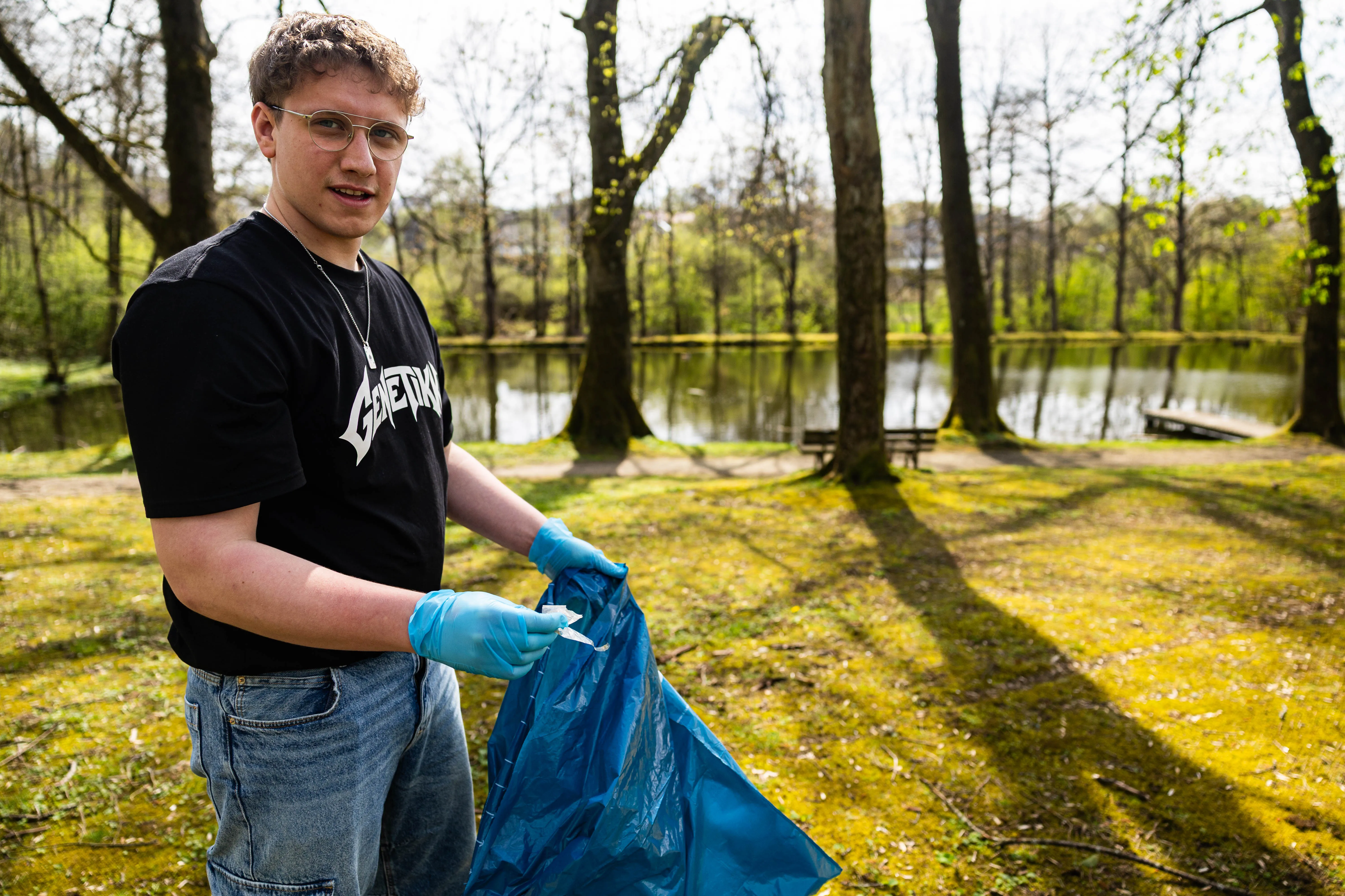 Ein junger Mann mit Brille und blauen Handschuhen hält einen vollen blauen Müllsack in einer Parklandschaft am Seeufer.