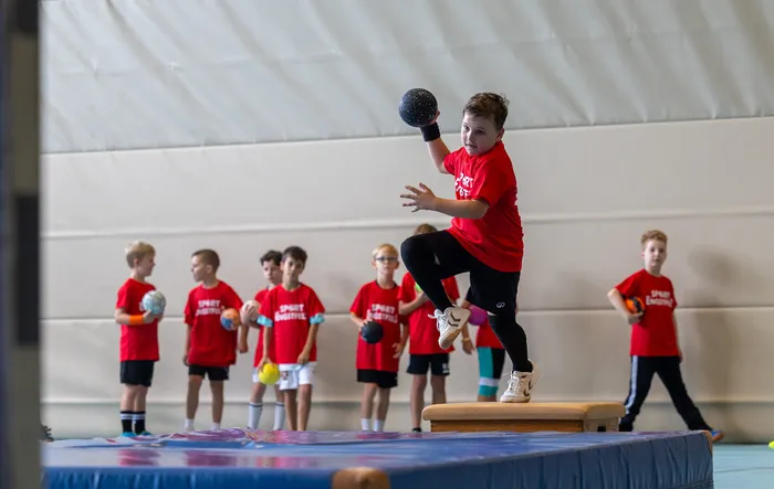 Ein Junge in einem roten T-Shirt springt mit einem schwarzen Ball in der Hand in einer Sporthalle hoch, während andere Kinder im Hintergrund zusehen.