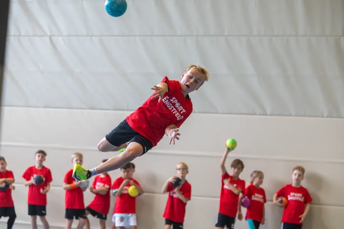 Ein Junge in einem roten T-Shirt springt in einer Sporthalle hoch in die Luft, während ein blauer Ball über ihm fliegt.