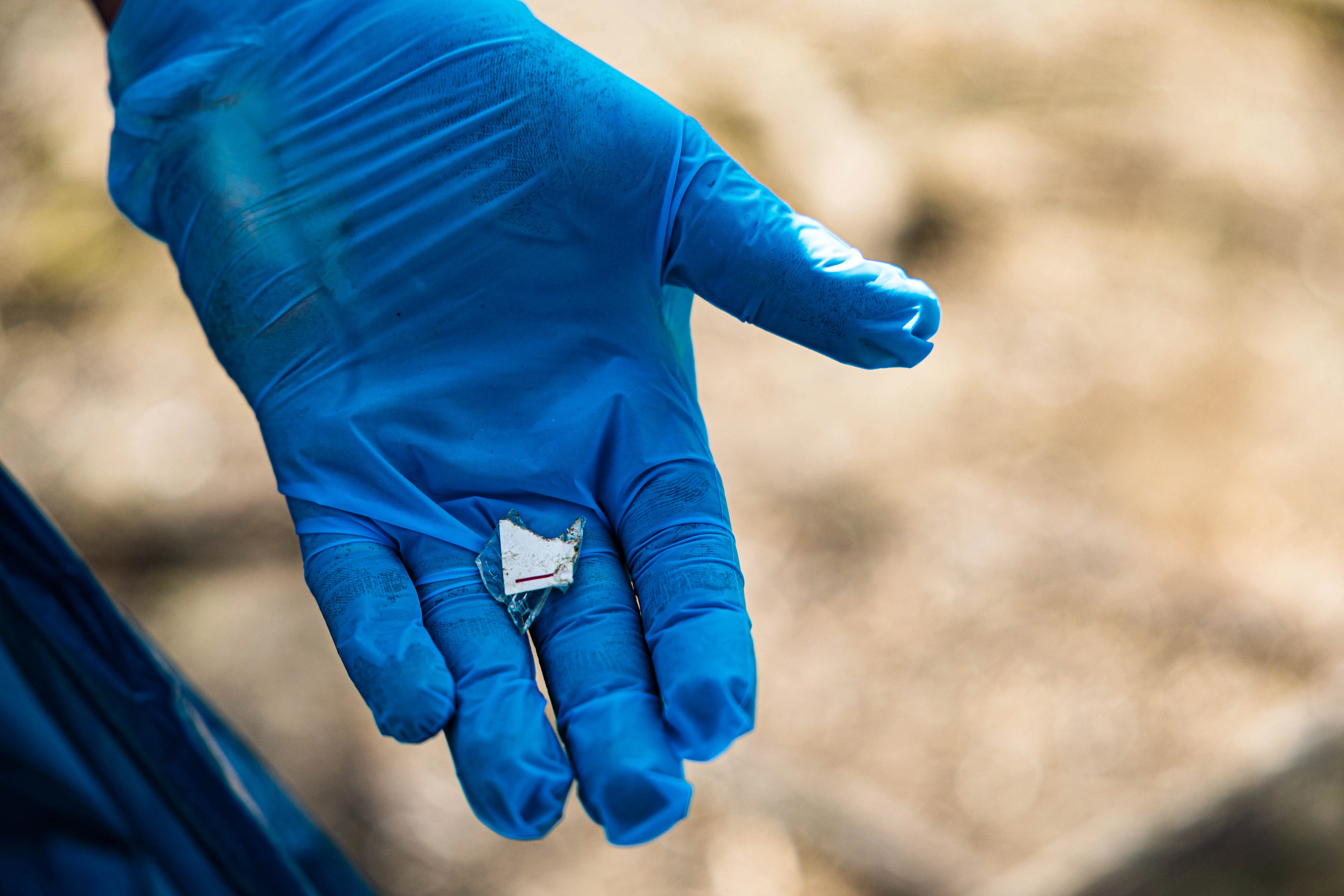 Blaue Handschuhe halten einen zerrissenen, silbernen Süßigkeitenpapierrest über unscharfem Untergrund.
