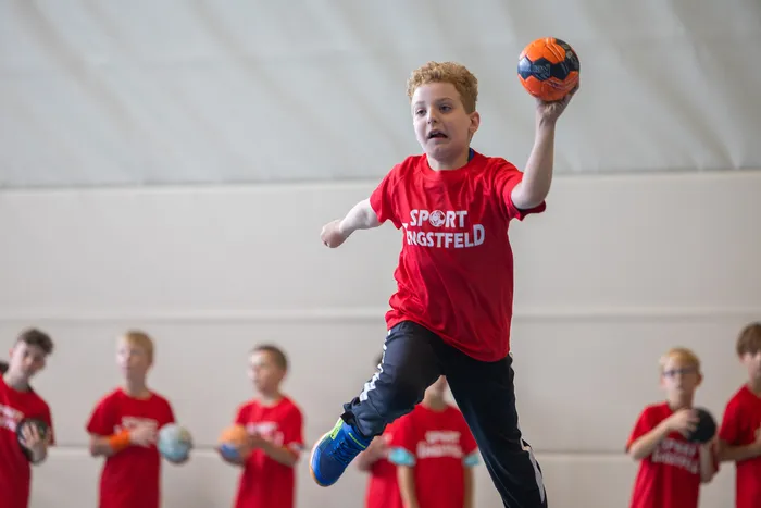 Ein Junge in einem roten T-Shirt springt in einer Sporthalle hoch und hält einen orange-schwarzen Ball zum Wurf bereit.
