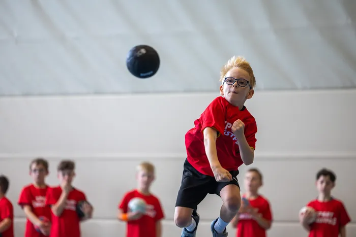 Ein Junge in einem roten T-Shirt springt nach einem schwarzen Ball, während andere Kinder im Hintergrund stehen.
