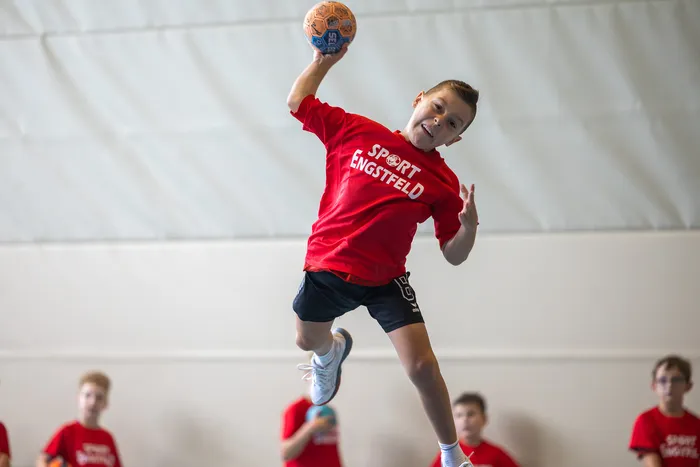 Ein Junge in einem roten T-Shirt mit der Aufschrift „SPORT ENGSTFELD“ springt in einer Sporthalle hoch in die Luft, um einen Handball zu werfen.