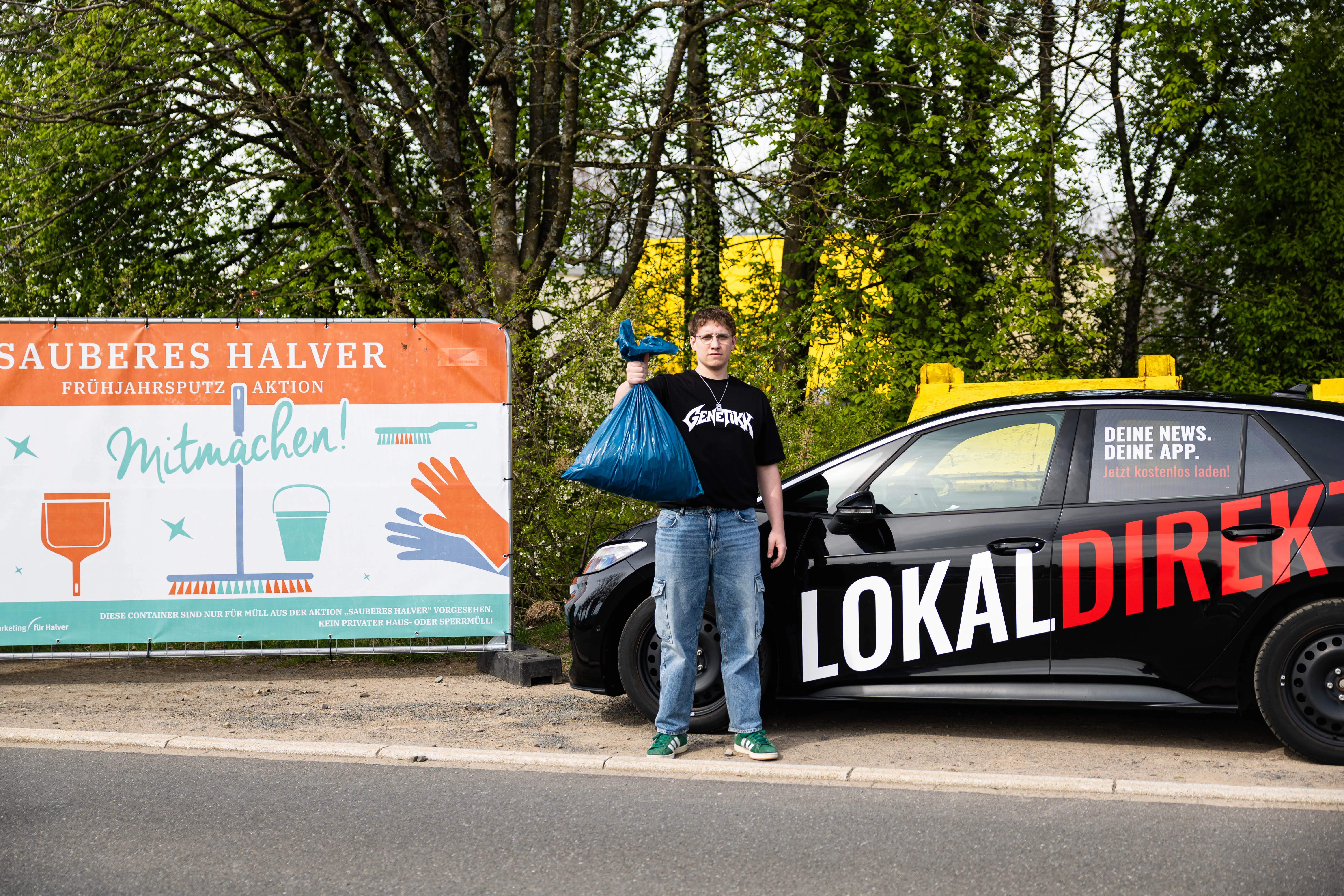 Junger Mann mit Müllsack steht neben einem Auto mit Aufschrift „LokalDirekt“ vor einem Plakat zur Müllsammelaktion „Sauberes Halver“.