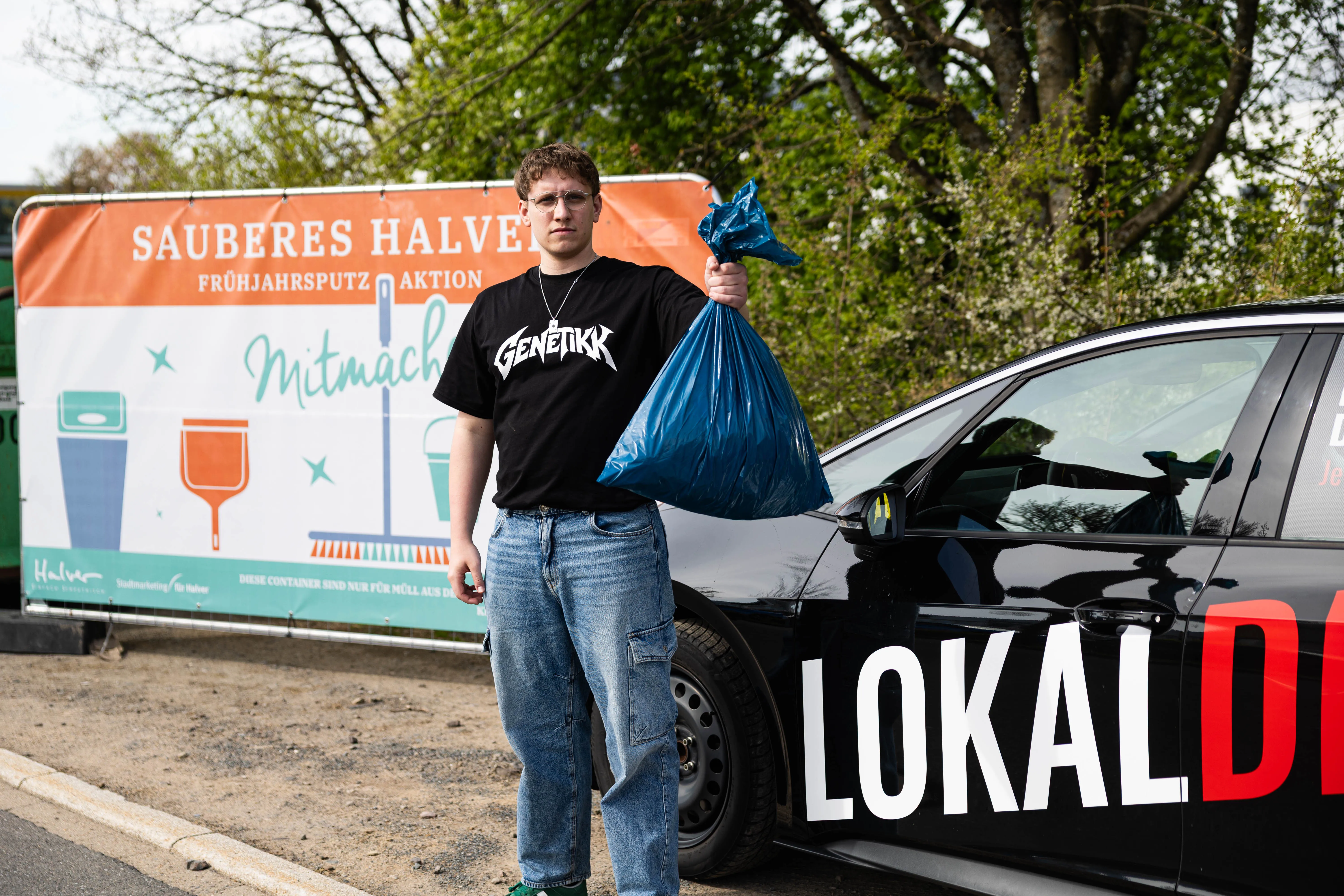 Ein junger Mann mit einem vollen blauen Müllsack steht vor einem Auto mit der Aufschrift LOKALDIREKT und einem Banner mit der Aufschrift SAUBERES HALVER FRÜHJAHRSPUTZ AKTION.