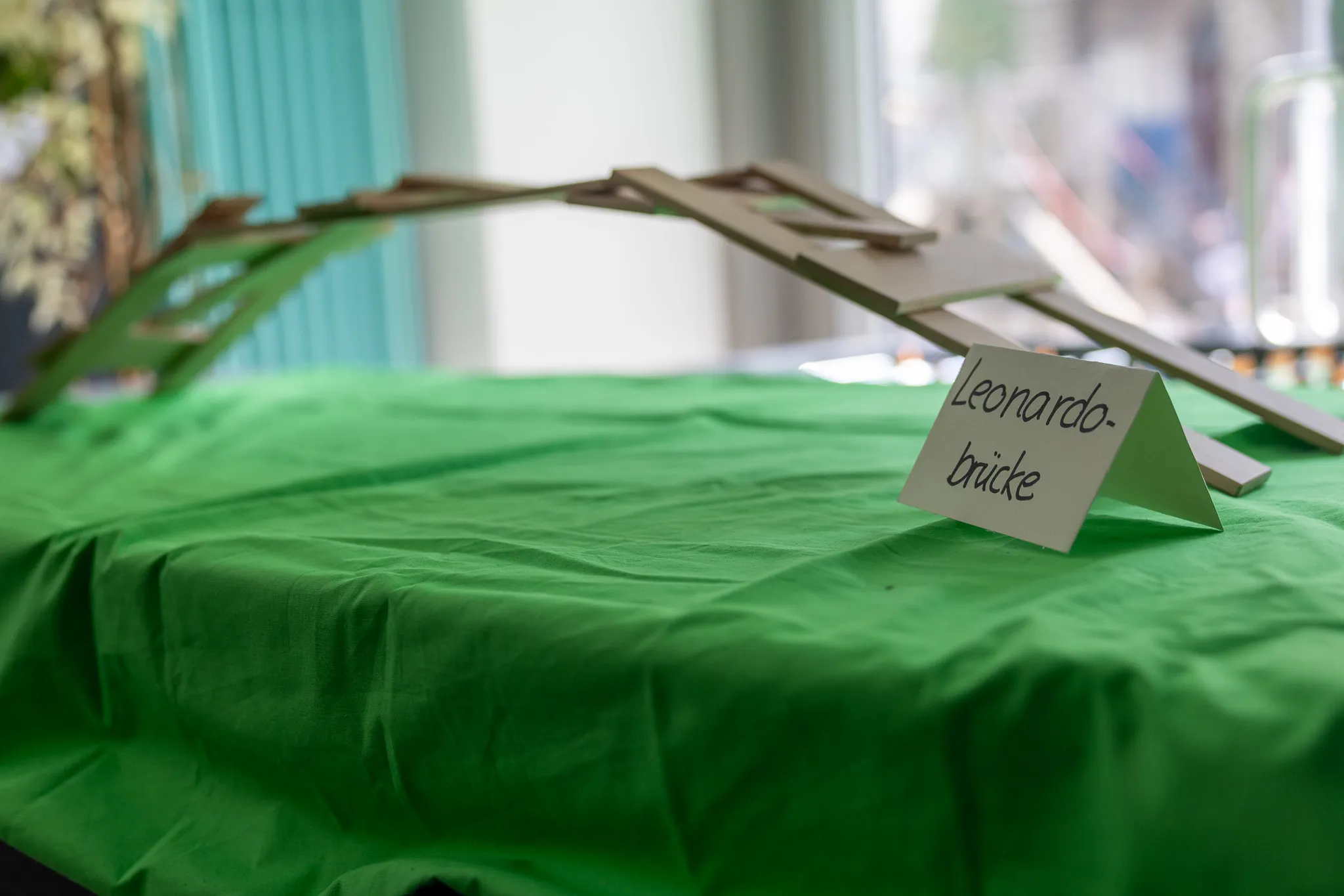 Modell einer Holzbrücke steht auf einer grünen Tischdecke vor einem verschwommenen Hintergrund. Ein Schild mit der Aufschrift „Leonardo-Brücke“ steht daneben.