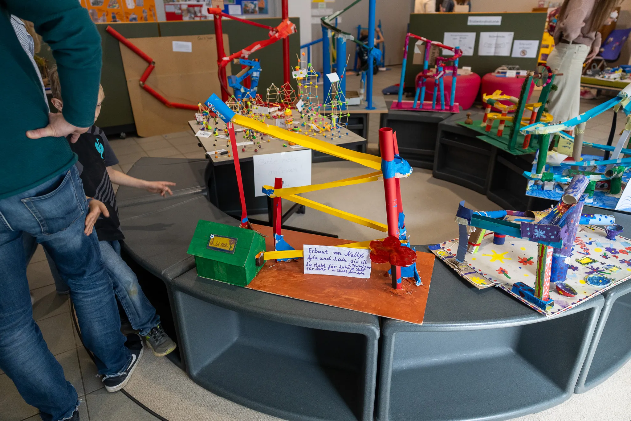Mehrere bunte Murmelbahnen aus Pappe und anderen Materialien stehen auf Tischen in einem Raum, vor denen eine Person steht und eine Bahn zeigt. Ein Schild neben einer Bahn trägt den Text: „Erstellt von Malte, Sofia und Luca aus der 3a“.