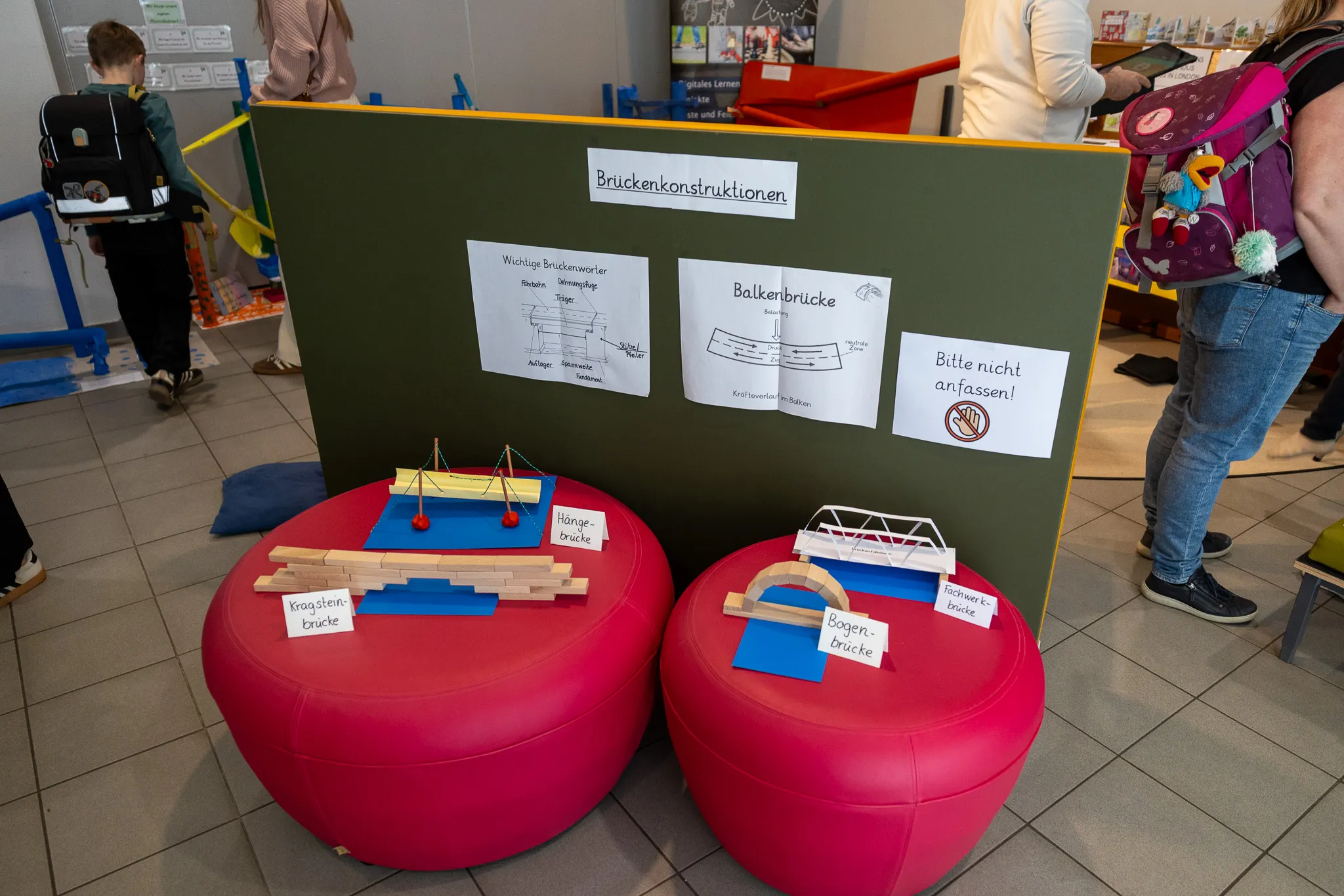Ein roter Tisch präsentiert verschiedene Brückenmodelle aus Holzstäben, darunter eine Hängebrücke und eine Bogenbrücke. Im Hintergrund eine Tafel mit Bauanleitungen und Warnhinweisen.