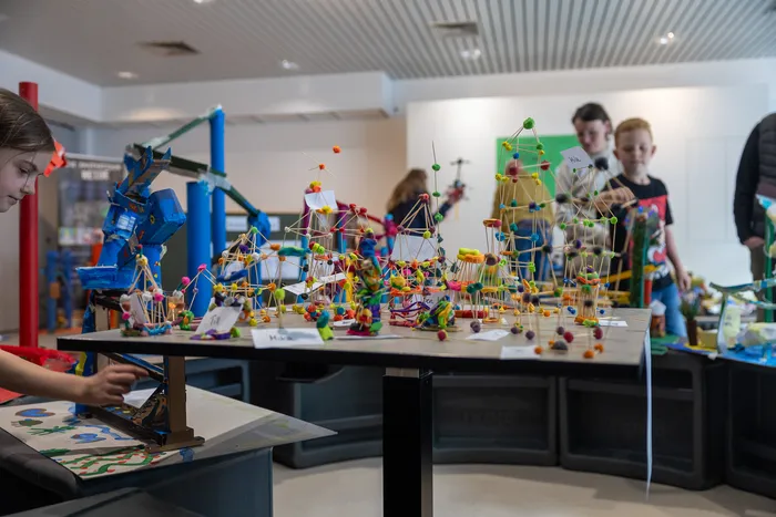 Schüler arbeiten an Modellen aus Bauklötzen und anderen Materialien auf einer schwarzen Tischreihe in einem hellen Raum.