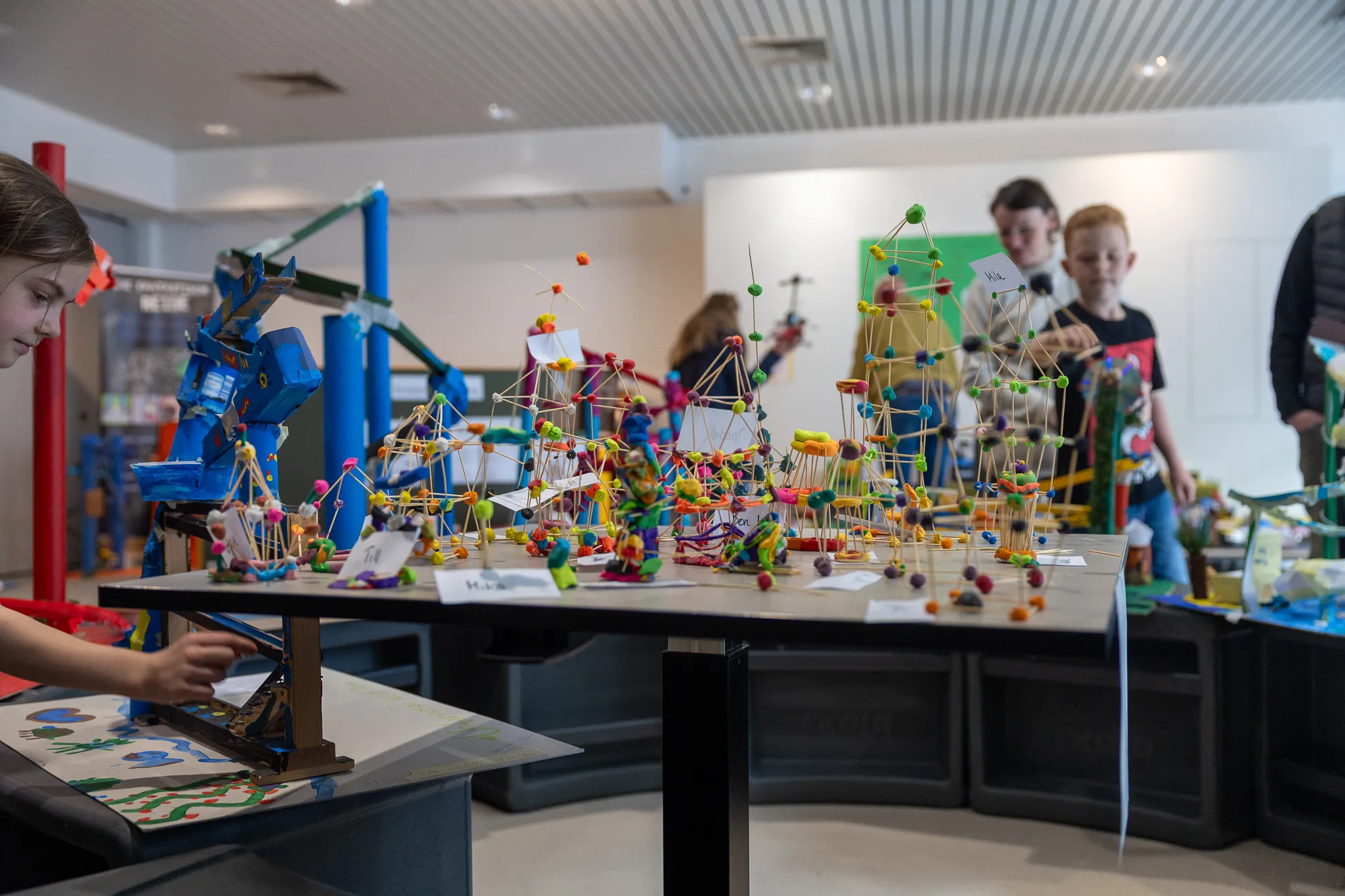 Schüler arbeiten an Modellen aus Bauklötzen und anderen Materialien auf einer schwarzen Tischreihe in einem hellen Raum.