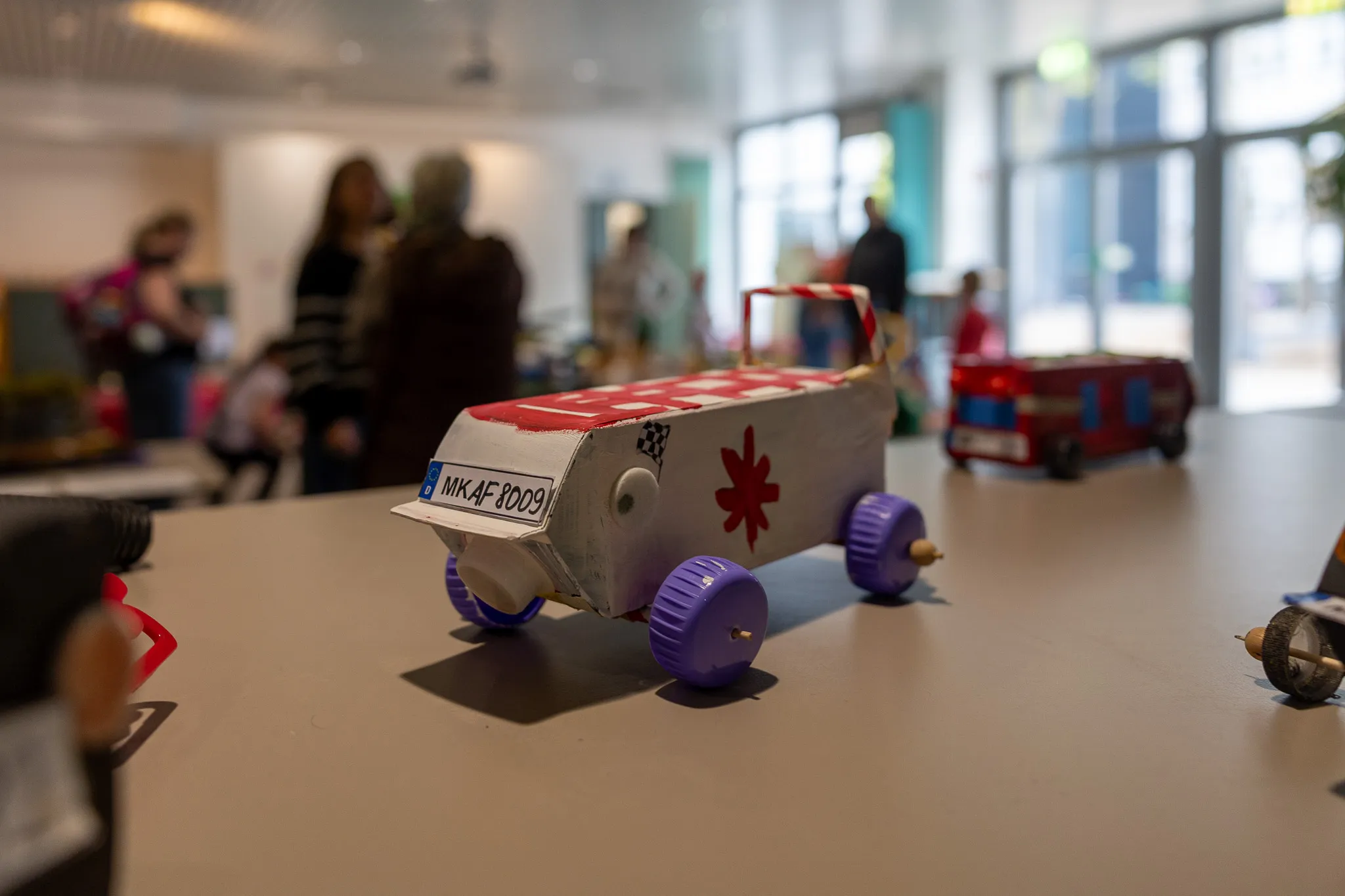 Handgefertigtes Auto aus Pappe mit rotem Kreuz auf der Seite steht auf einem Tisch, im Hintergrund verschwommene Personen.
