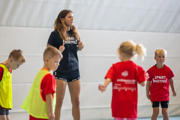 Eine Frau in einem schwarzen T-Shirt mit der Aufschrift „SPORT ENGSTFELD“ steht vor einer Gruppe von Kindern in einer Sporthalle.