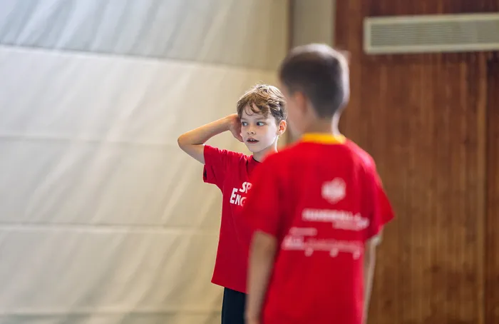 Ein Junge in einem roten T-Shirt blickt zur Seite und berührt seinen Kopf, während ein zweiter Junge im Vordergrund unscharf zu sehen ist.