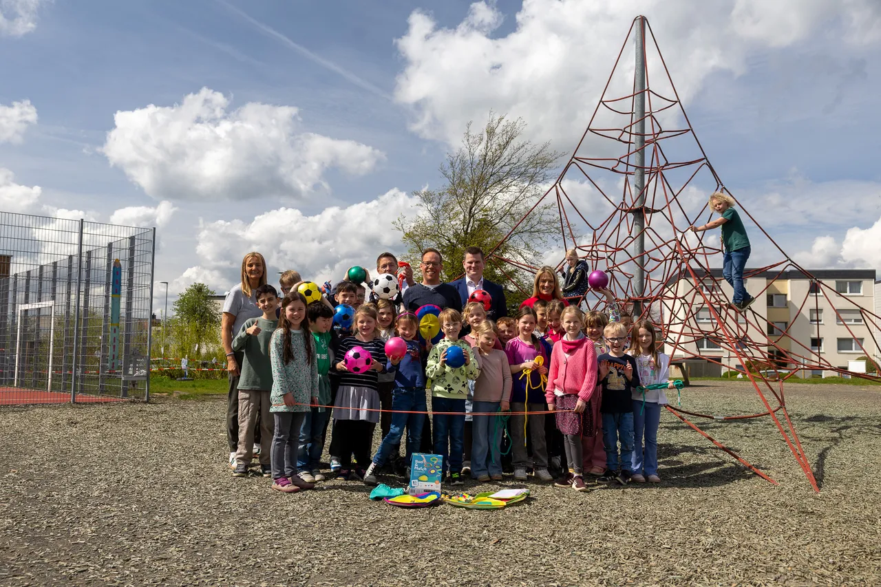Eine Gruppe von Kindern und Lehrern steht vor einem großen Klettergerüst auf einem Kiesplatz. Im Hintergrund ein Fußballfeld und Wohngebäude unter einem bewölkten Himmel.