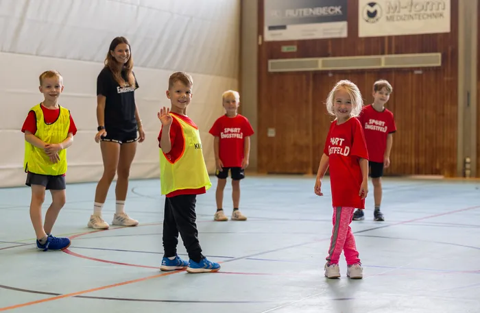 Kinder in roten T-Shirts und gelben Sportwesten sowie eine junge Frau stehen lächelnd in einer Sporthalle.