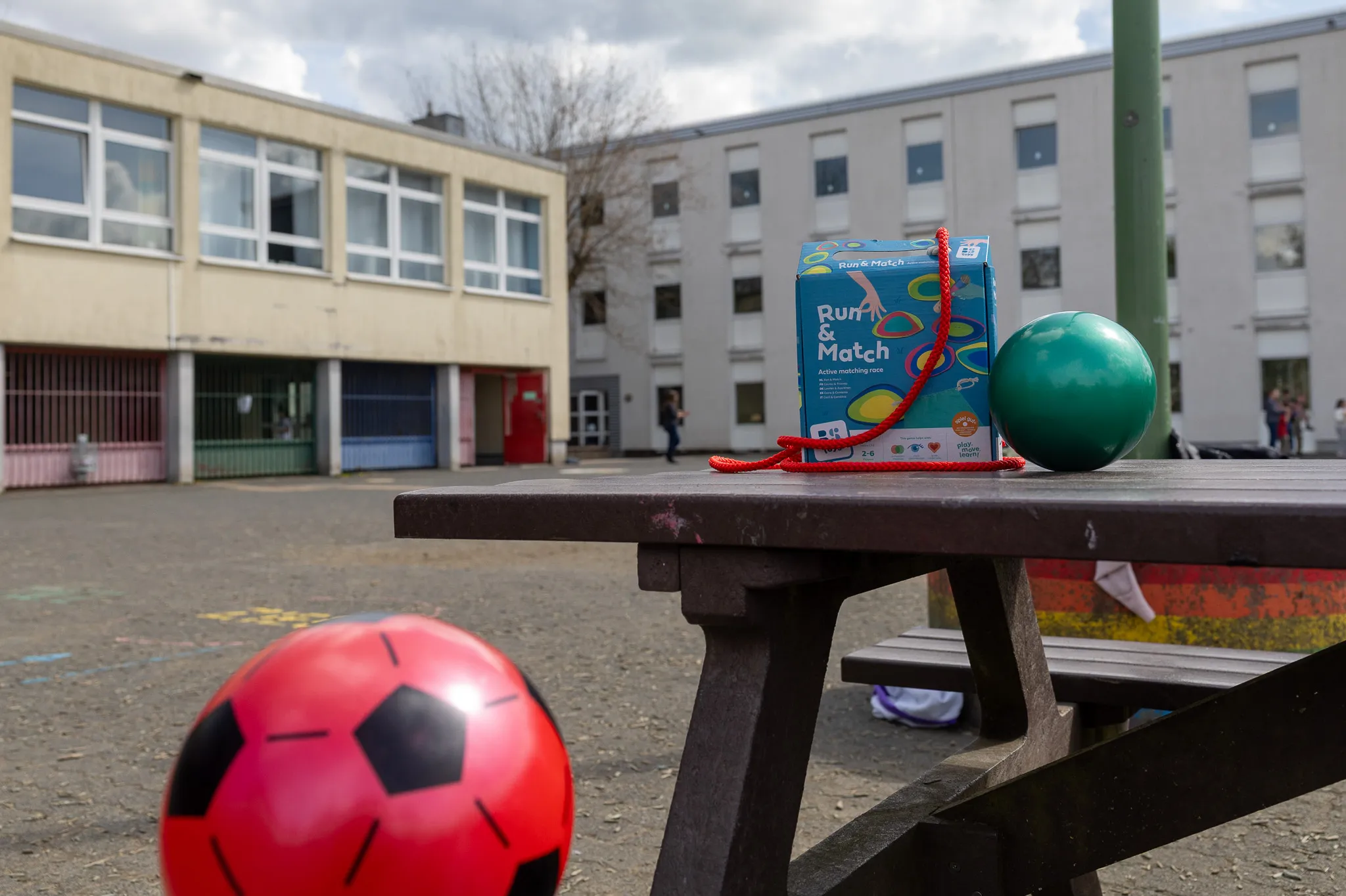 Spielzeug auf einem Picknicktisch im Schulhof: ein Fußball, ein grüner Ball und eine Spielzeugbox mit dem Aufdruck „Run & Match“.