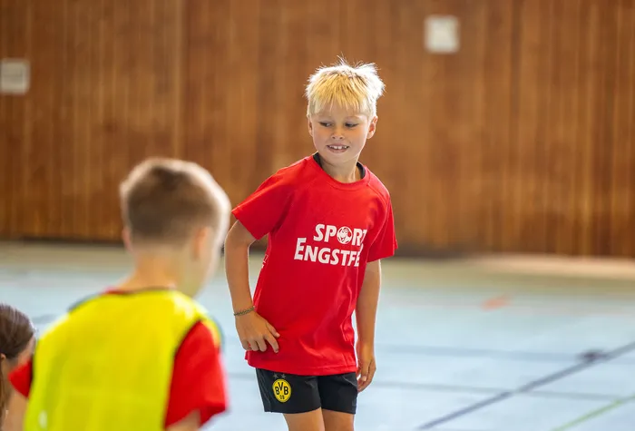 Ein lächelnder Junge mit blonden Haaren trägt ein rotes T-Shirt mit der Aufschrift „SPORT ENGSTEF“ in einer Sporthalle.