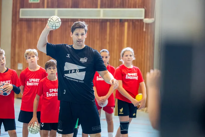 Ein Mann in schwarzem Sportshirt bereitet einen Ballwurf vor, während Kinder in roten T-Shirts im Hintergrund zuschauen.