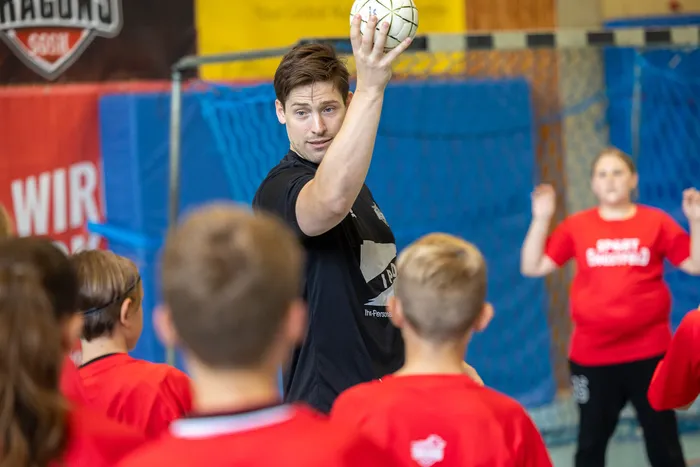 Ein Mann hält einen Handball hoch und zeigt ihn einer Gruppe von Kindern in roten T-Shirts in einer Sporthalle.