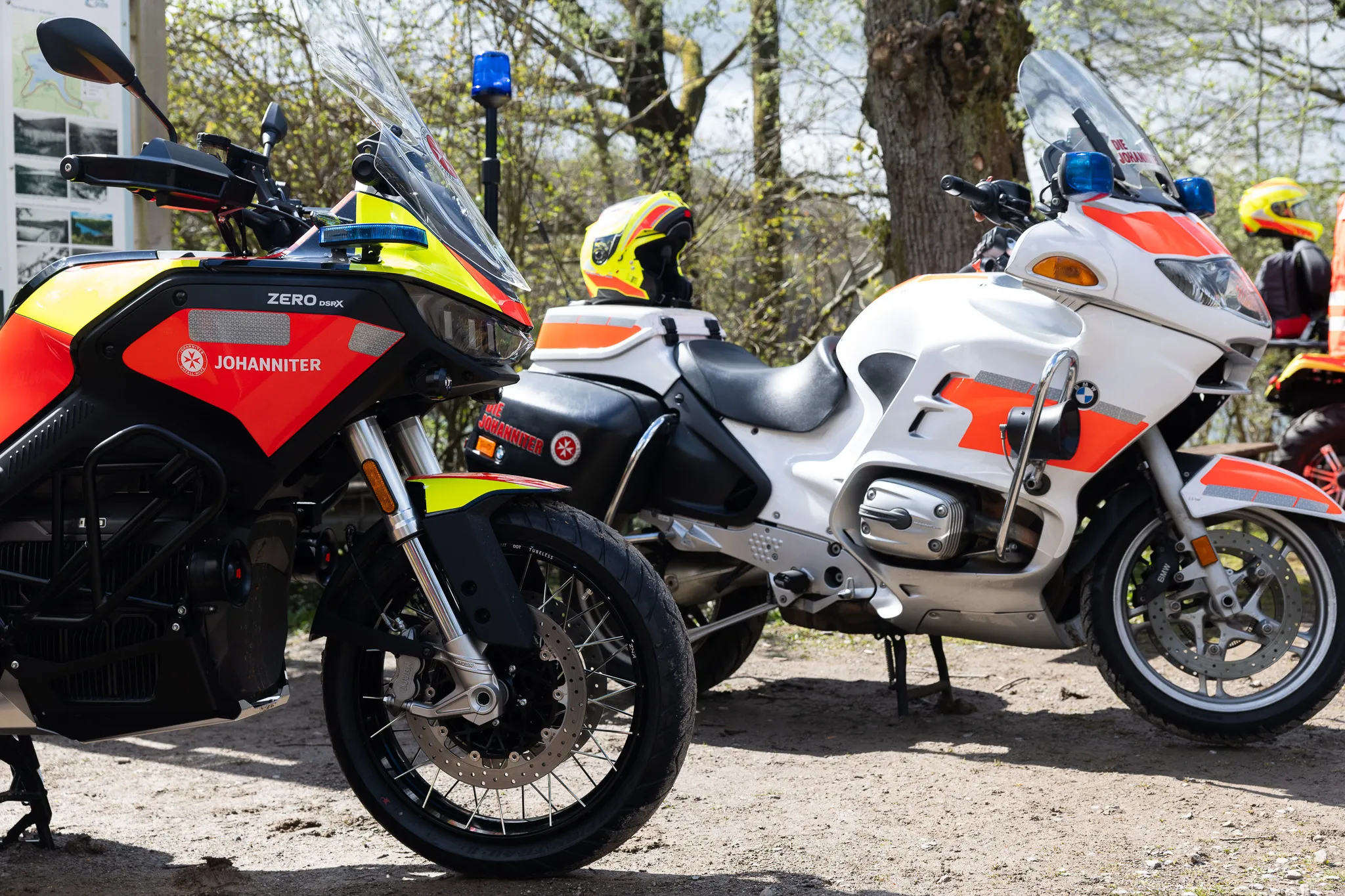 Zwei Motorräder, ein schwarzes Zero DSX und eine weiße BMW, mit Kennzeichnung des Johanniter-Unfallhilfsdienstes stehen nebeneinander auf einem Kiesweg. Im Hintergrund sind Helme und weitere Einsatzkräfte zu sehen.