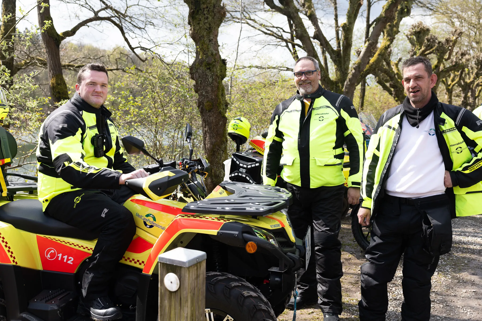 Vier Männer in fluoreszierenden Warnwesten stehen neben einem gelben Quad mit Blaulicht und dem Aufdruck „MedCare Professional“ im Wald.