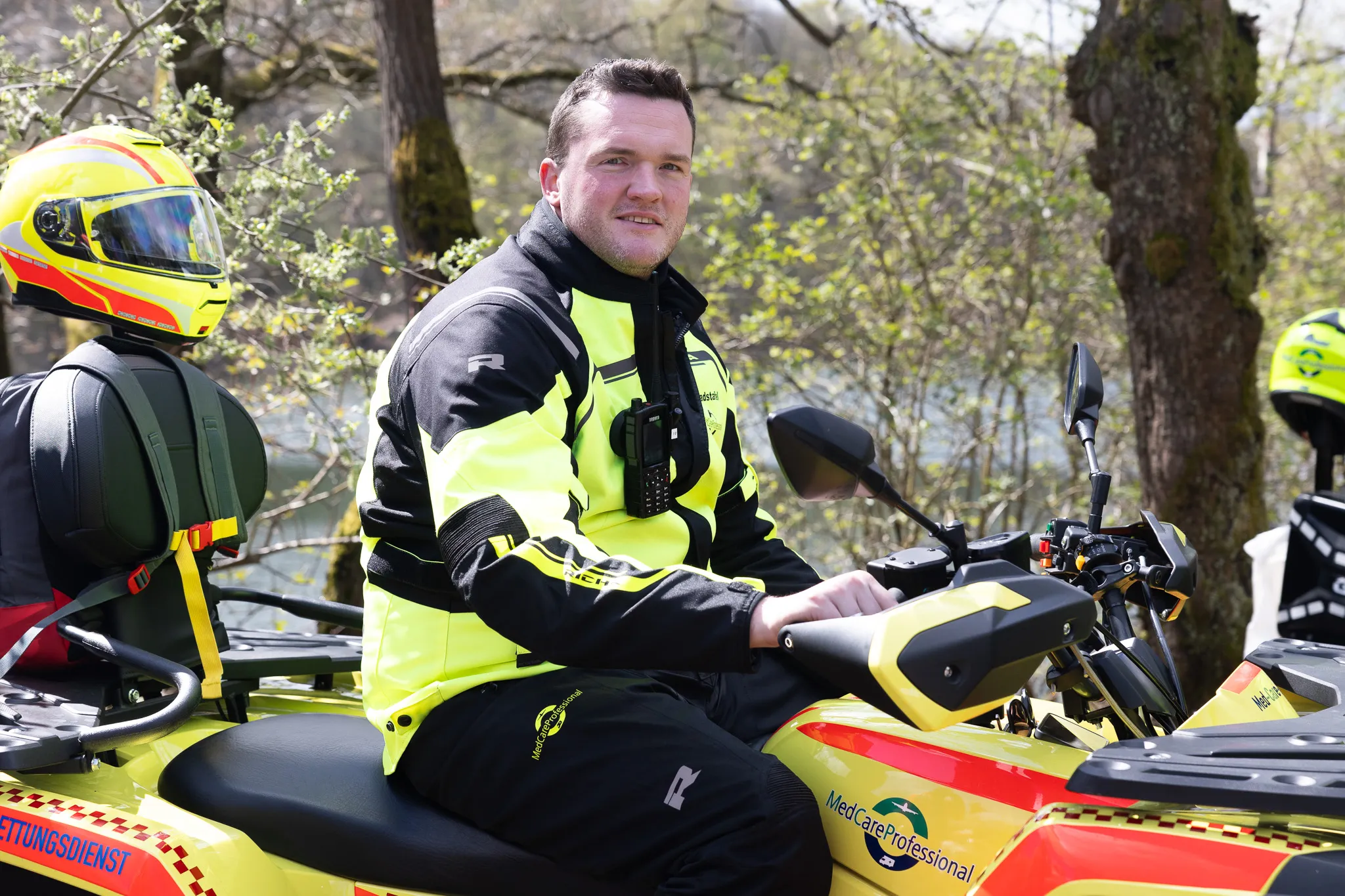 Ein Rettungsdienstmitarbeiter in gelber und schwarzer Schutzkleidung sitzt auf einem gelben Quad. Im Hintergrund sind Bäume und weitere Einsatzkräfte zu sehen.