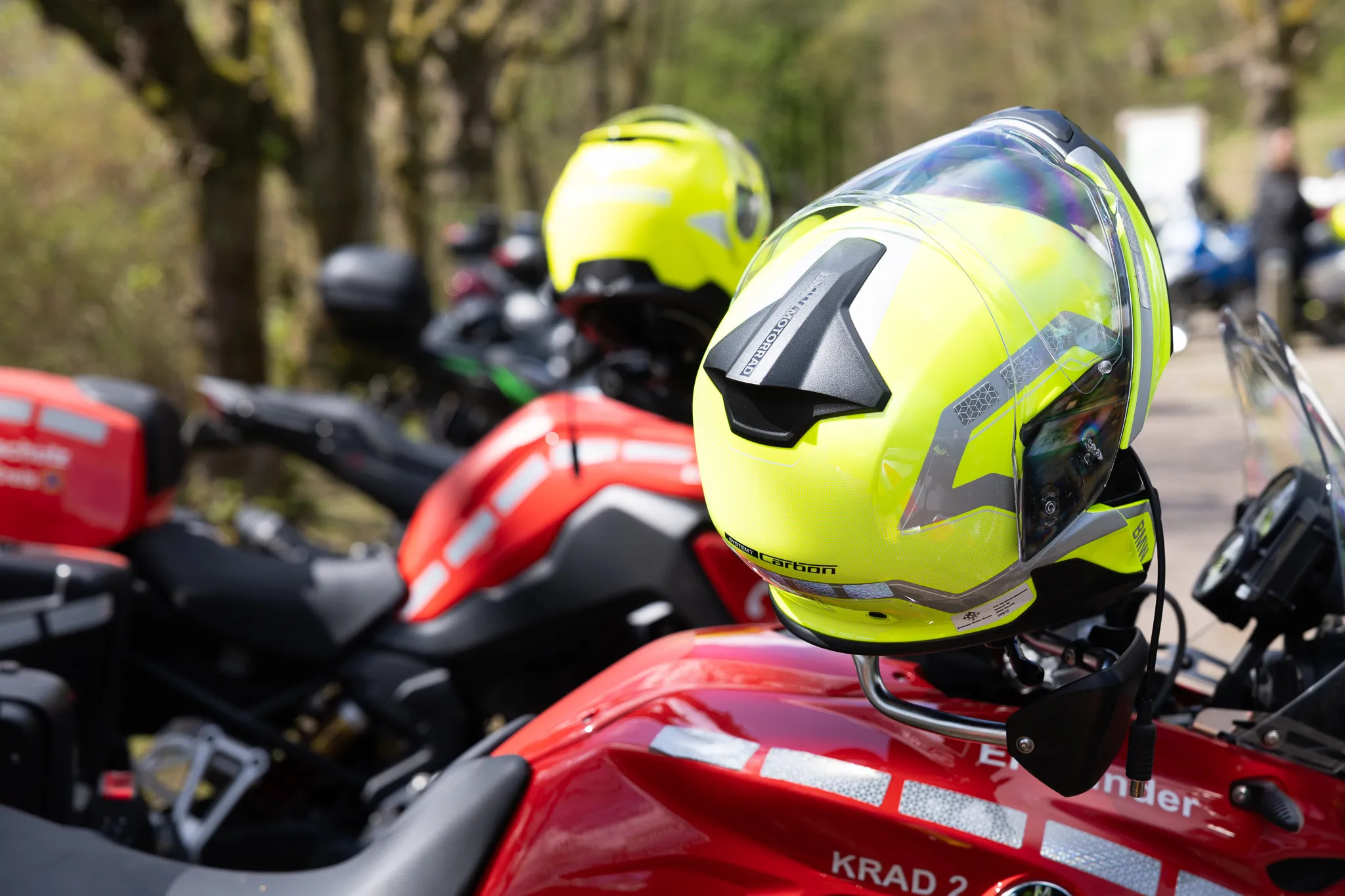 Neon-gelber Motorradhelm mit Visier auf einem roten Motorrad, im Hintergrund weitere Motorräder und Bäume.