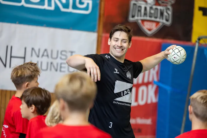 Ein lächelnder Mann in schwarzem Sportshirt hält einen Handball hoch, umgeben von Kindern in roten T-Shirts in einer Sporthalle.