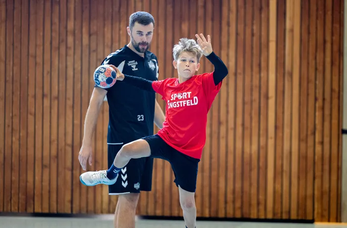 Ein Junge in einem roten T-Shirt springt mit einem Handball in der Hand, während ein Mann in einem schwarzen Sportshirt hinter ihm steht.