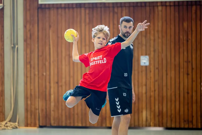 Ein Junge in einem roten T-Shirt springt mit einem gelben Handball in der Hand hoch, um ihn zu werfen.